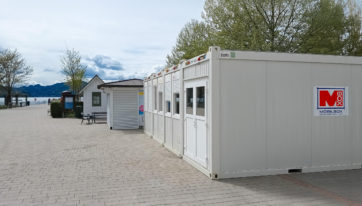 Ein Mobilbox Bürocontainer von außen, platziert am Ufer des Wörthersees in Klagenfurt. Die Container haben Fenster mit geschlossenen Rollläden und eine Eingangstür. Das Mobilbox-Logo ist an den Containern sichtbar. Im Hintergrund sind der See und eine bergige Landschaft zu erkennen.