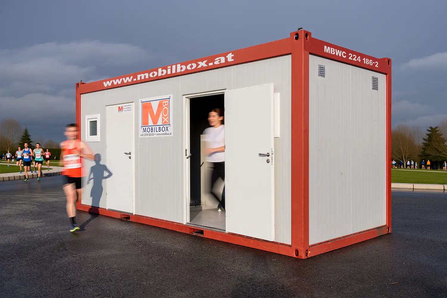 WC-Container mit geöffneter Tür während Marathonläufer vorbeilaufen bei einer Sportveranstaltung.