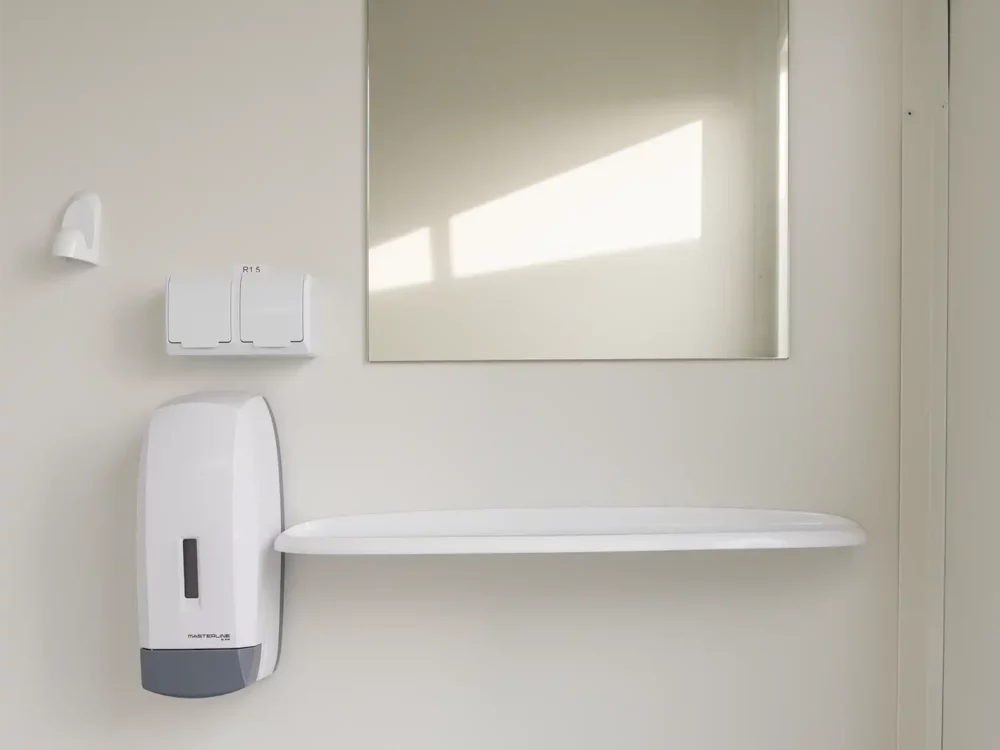 Detailansicht eines WC-Containers mit Blick auf eine Ablage, Spiegel, Seifenspender sowie einen Klebehaken an der Wand.
