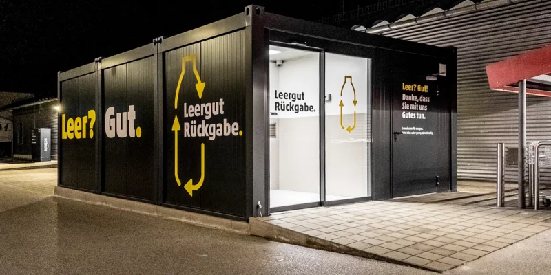 Container für Pfandflaschen-Rückgabe Automaten aufgestellt vor einem Supermarkt mit einer Schiebetür sowie Beklebung.